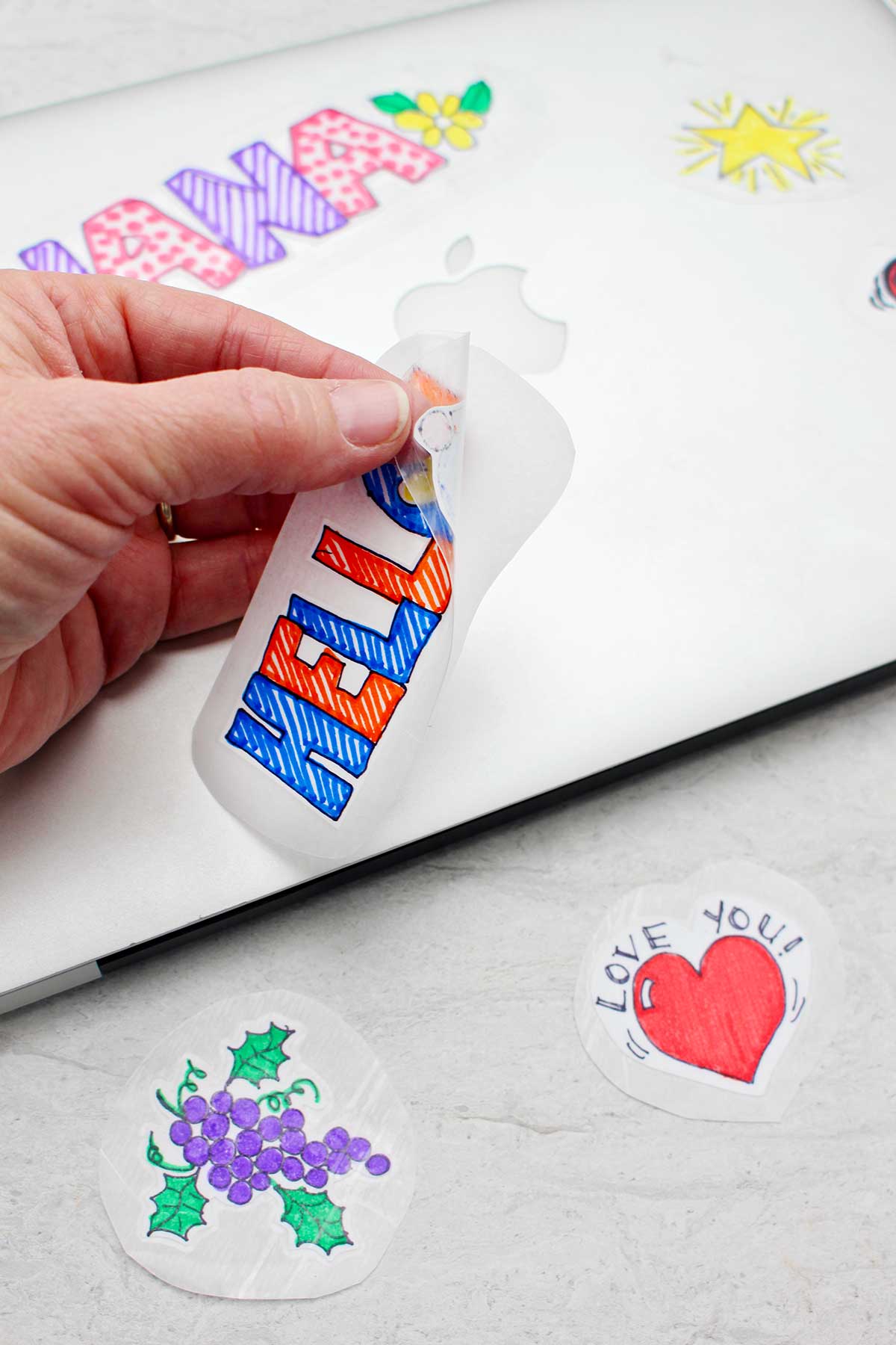 Hand peeling back a sticker that says "HELLO" with silver laptop and stickers in the background.