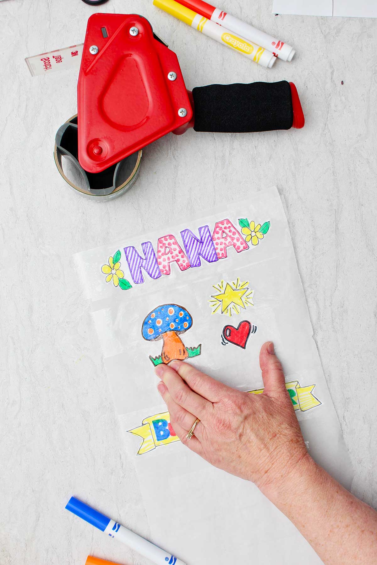 Hand securing tape over stickers on parchment paper with tape dispenser and markers near by.