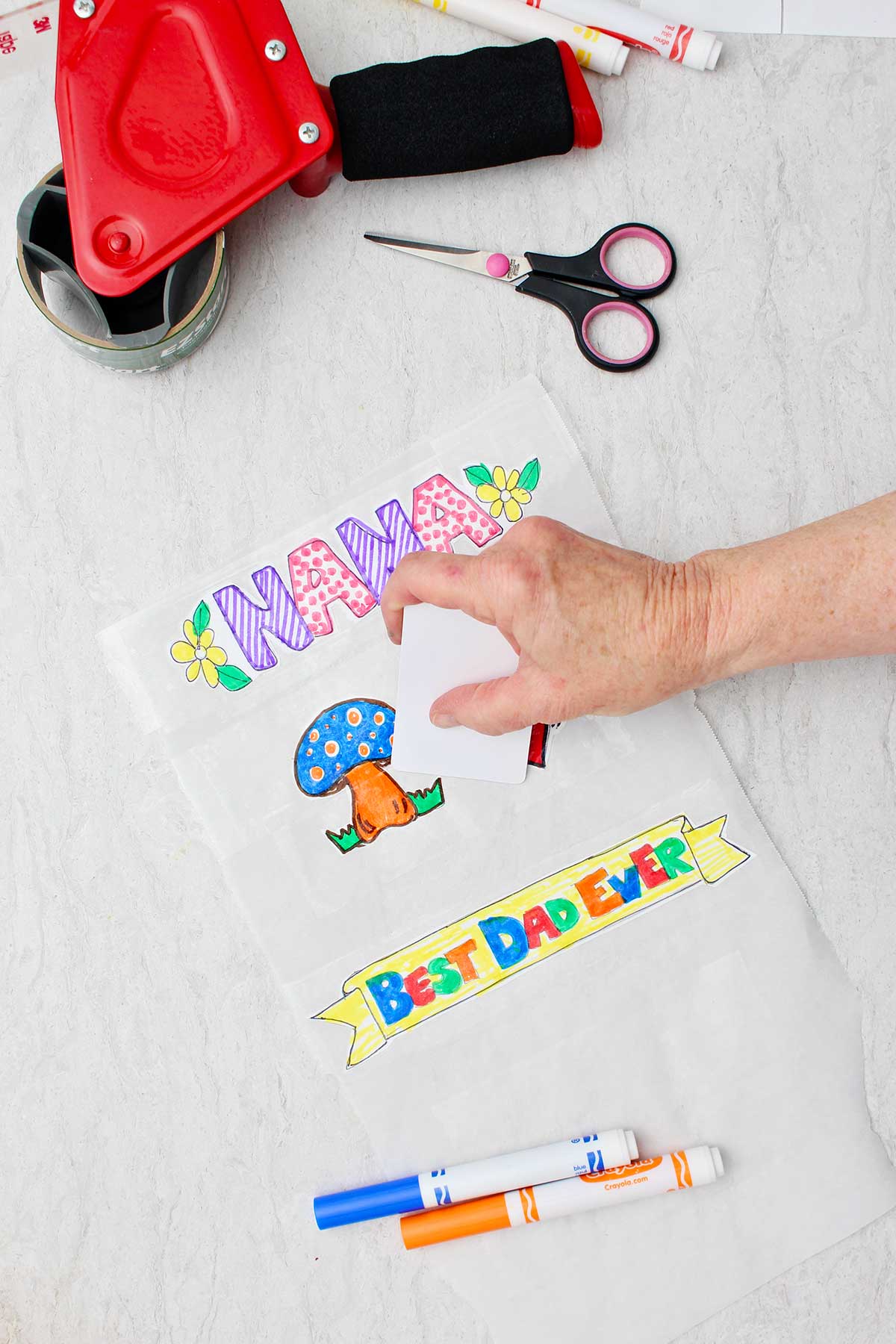 Hand pressing over the tape and parchment paper stickers with the edge of a plastic card with tape dispenser, scissors and markers near by.