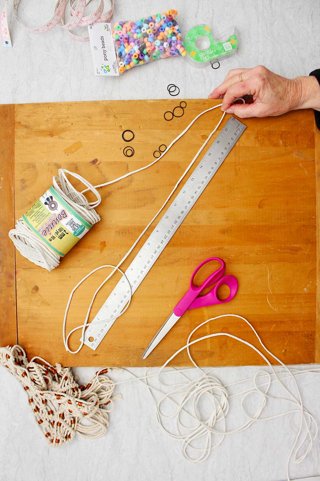 Hand measuring craft cord with a metal ruler on a wooden board with other supplies near by.