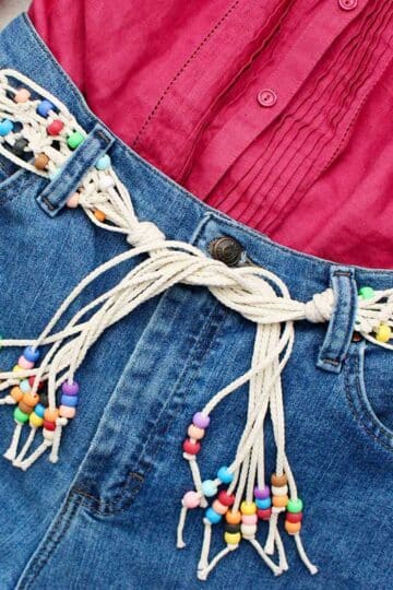 Macrame belt with colorful beads threaded through denim belt loops with a pink shirt on top.