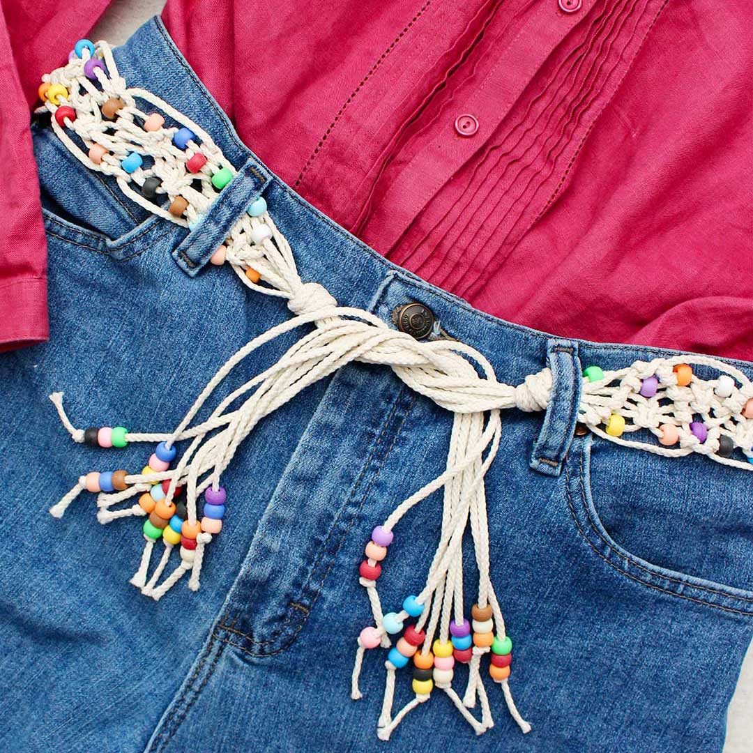 Macrame belt with colorful beads threaded through denim belt loops with a pink shirt on top.