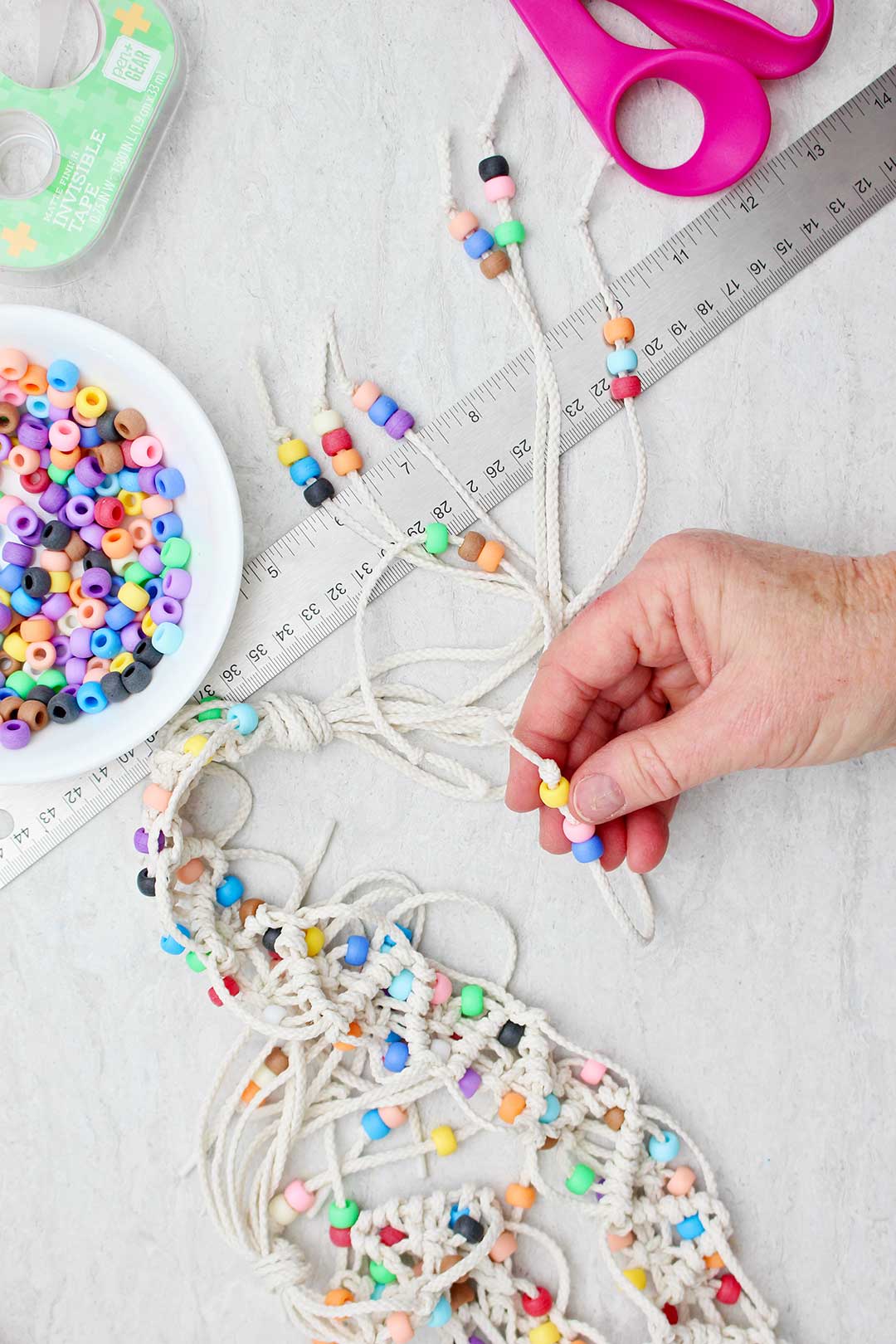 Hand holding up the beaded tassel end of a completed macrame belt with supplies near by.