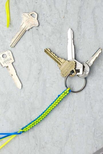 A blue and yellow completed boondoggle keychain with three keys attached with supplies near by.