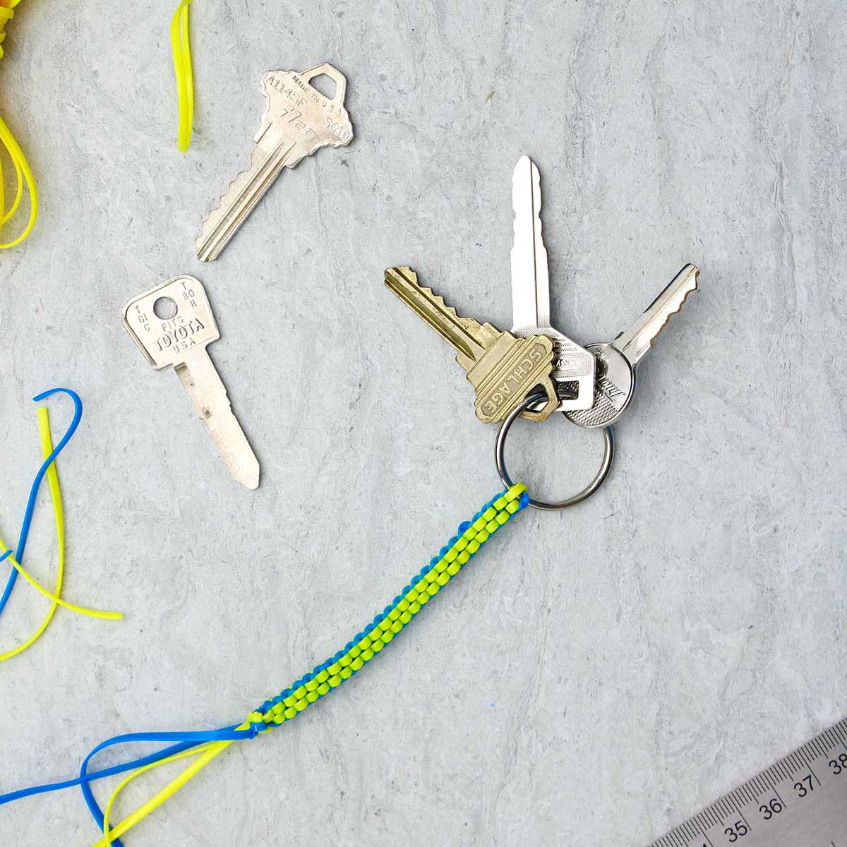 A blue and yellow completed boondoggle keychain with three keys attached with supplies near by.