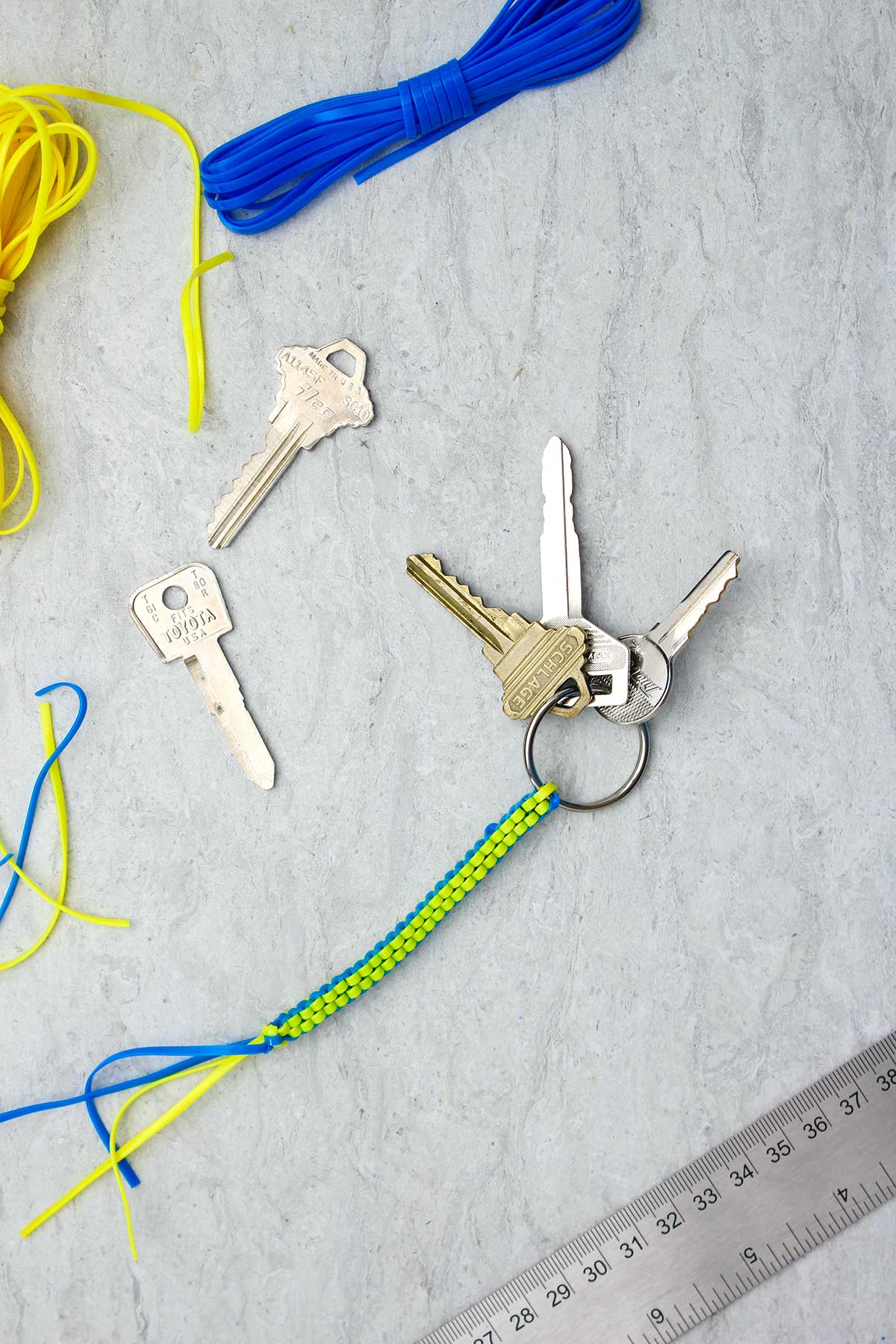 A blue and yellow completed boondoggle keychain with three keys attached with supplies near by.