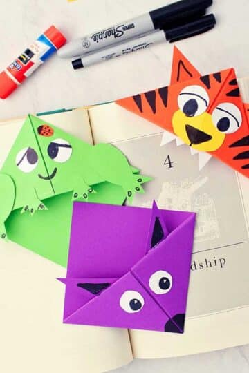 Three completed animal origami bookmarks in opened book with scraps of paper, sharpie markers and other supplies near by.