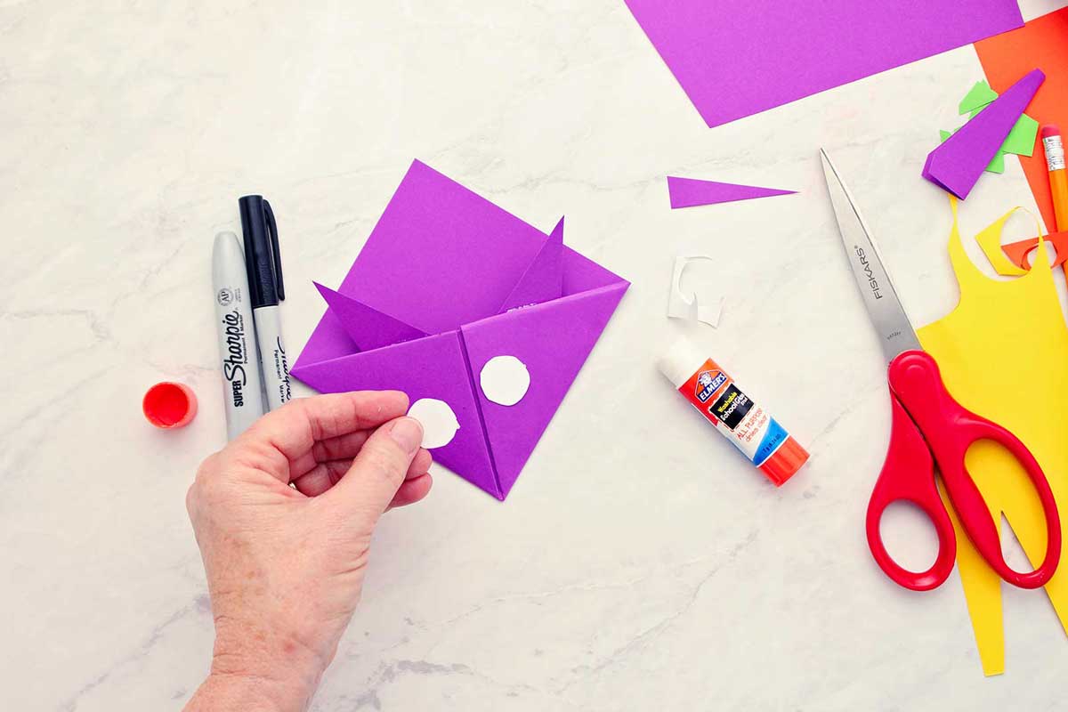 Hand placing white circles on purple origami fox bookmark for eyes.