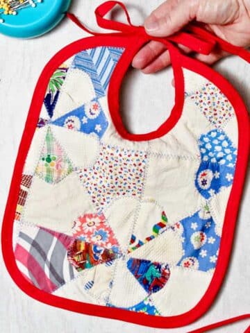 Hand holding completed quilted baby bib with scraps of fabric and a magnet of straight pins near by.