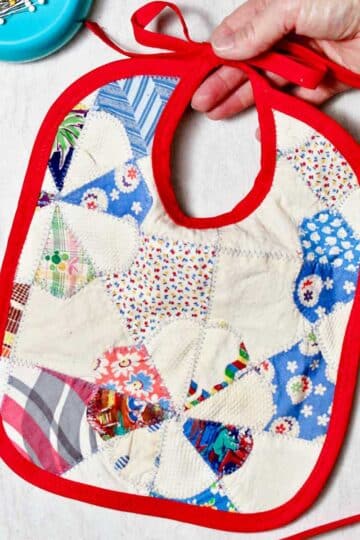 Hand holding completed quilted baby bib with scraps of fabric and a magnet of straight pins near by.