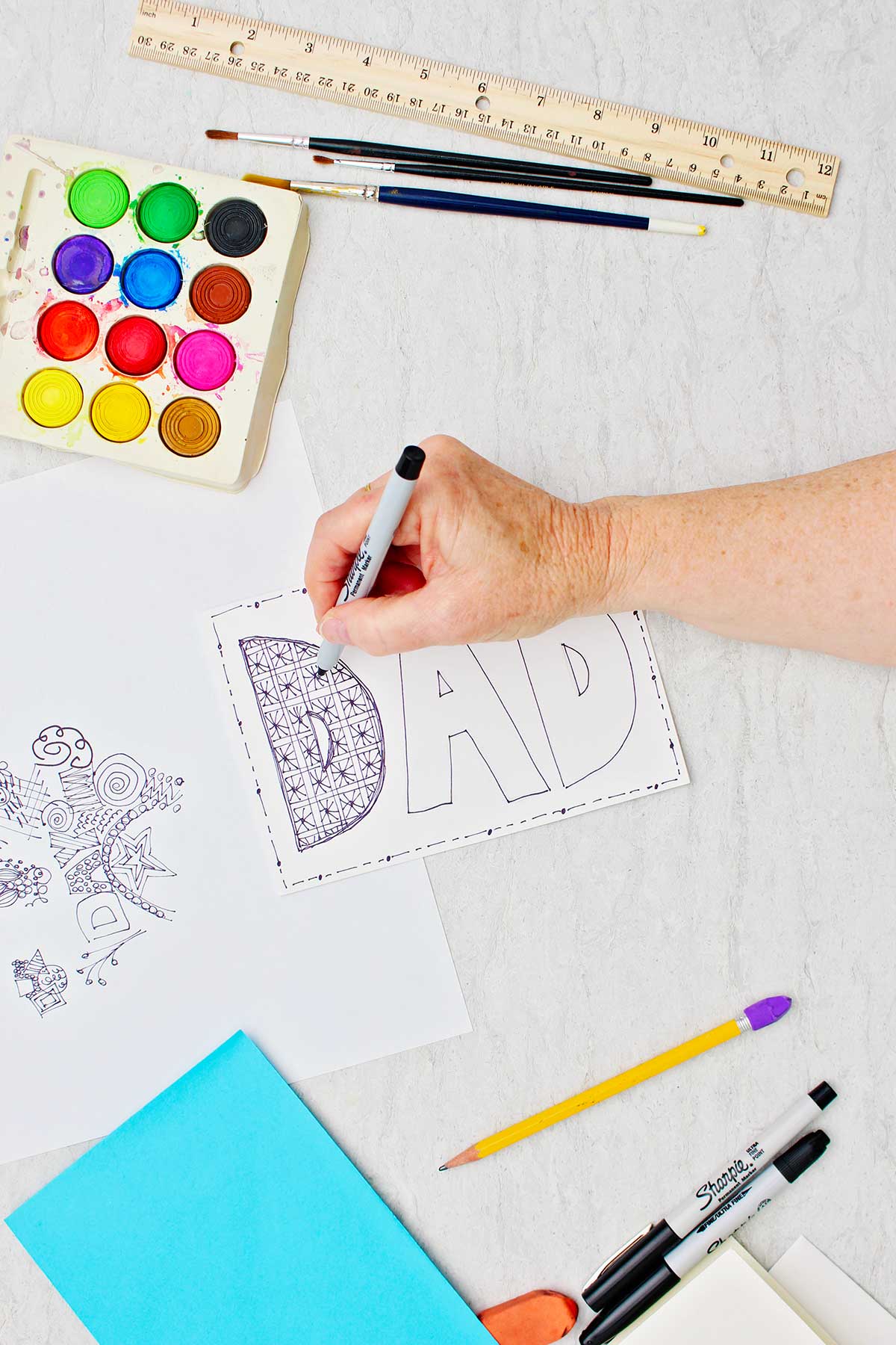 Hand doodling block letters in the word "DAD" on card with sharpie with other supplies near by.