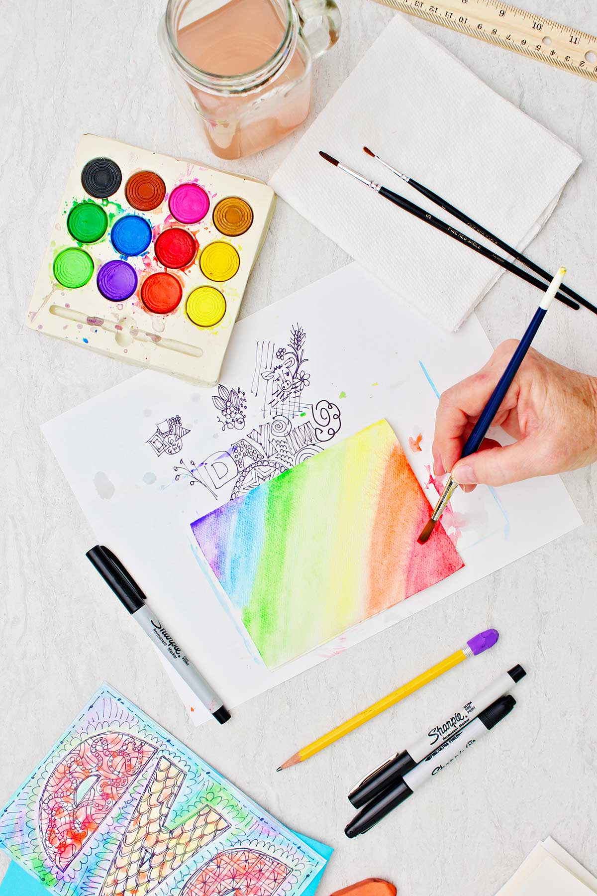 Hand painting card in a diagonal rainbow pattern with watercolors with other supplies near by.