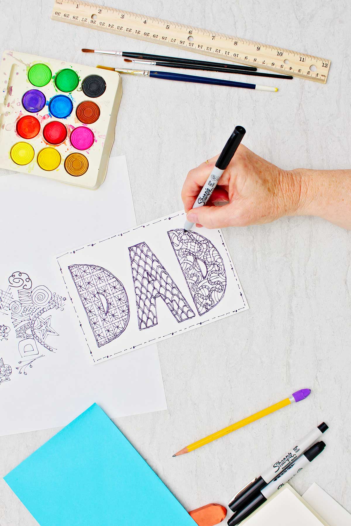 Hand doodling block letters in the word "DAD" on card with sharpie with other supplies near by.