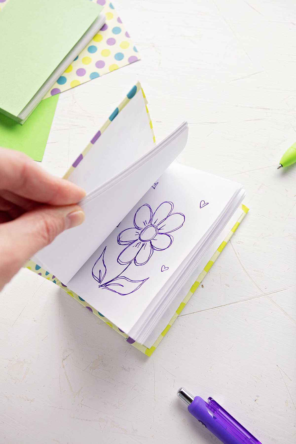 A DIY miniature journal with a flower picture drawn on one of the pages, scrapbook paper and pens in the background.