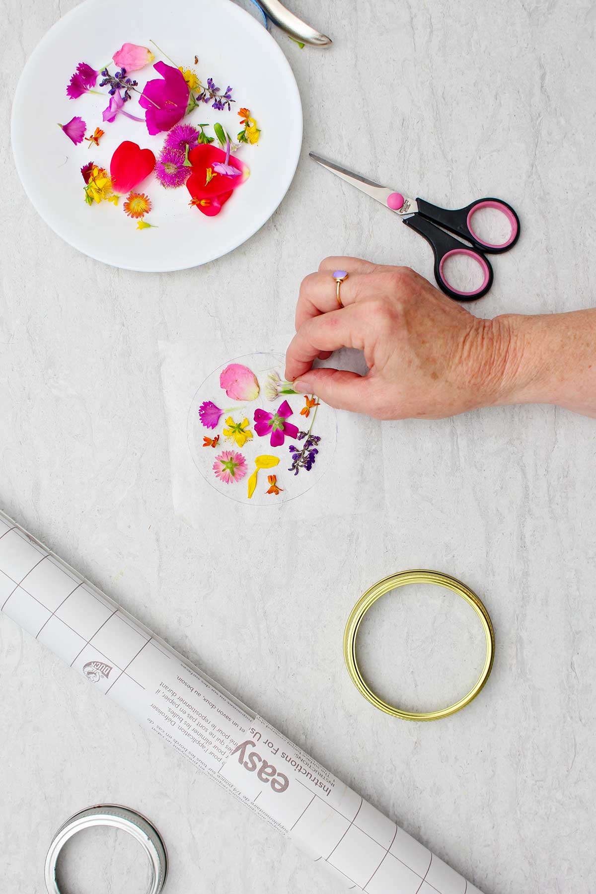 Person placing petals on clear laminate paper with other supplies near by.