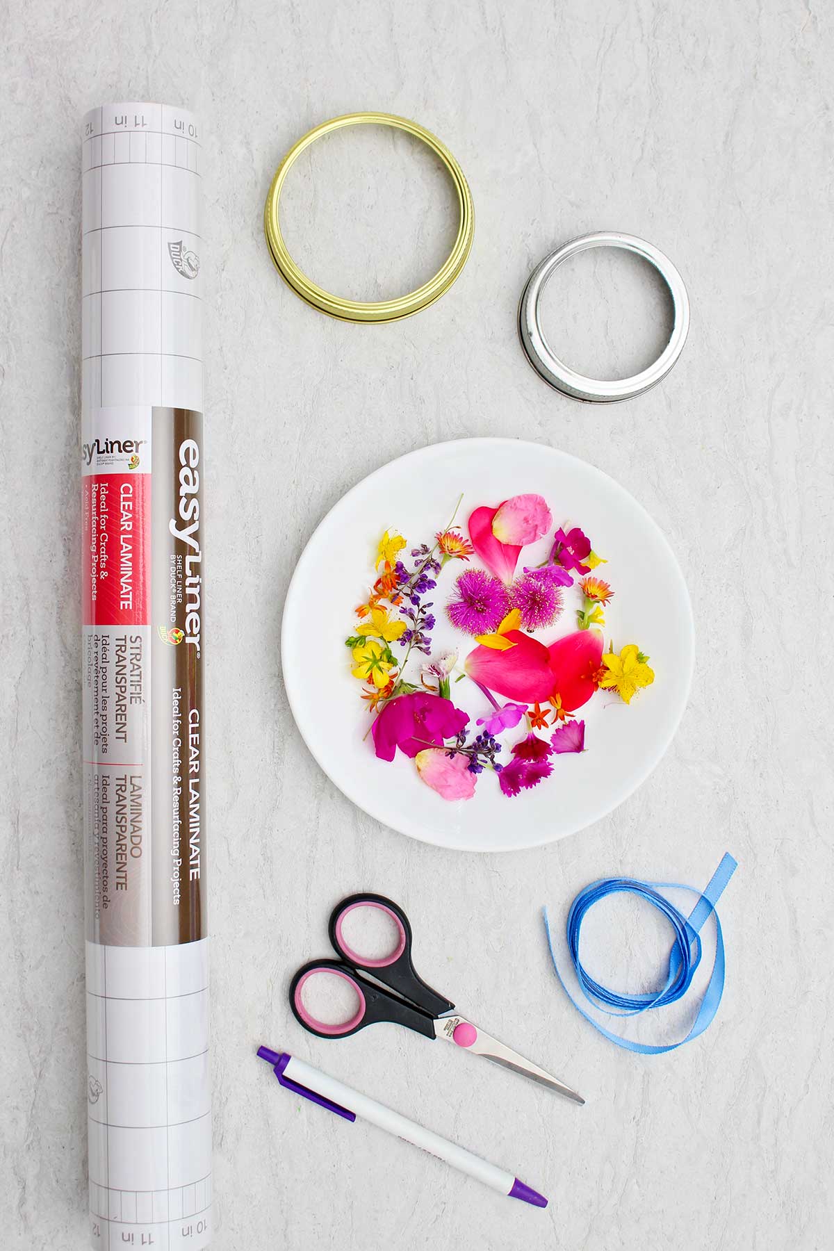 Clear laminate paper, mason jar lids, a plate of flower petals, blue ribbon, scissors and a pen.