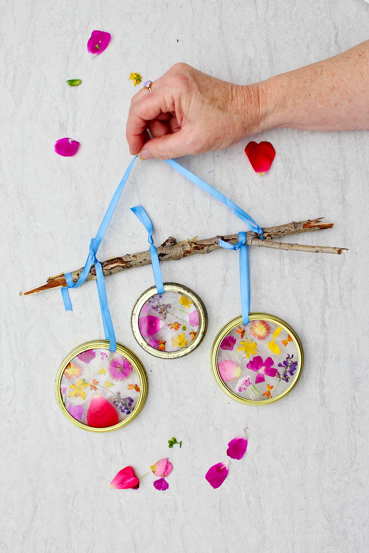 Completed Garden Suncatchers with pressed flowers made with a mason jar lid and hanging from a twig with blue ribbon against marble counter top.