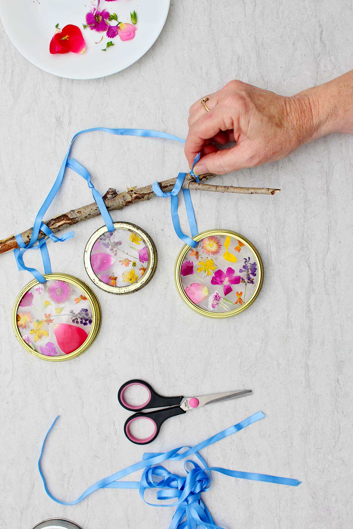 Person stringing blue ribbon around a twig to hang sun catchers with ribbon, scissors and flower petals near by.