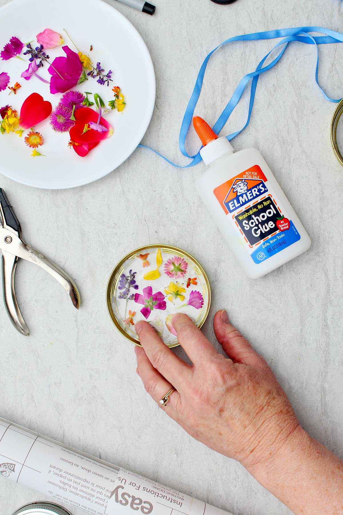 A plate of flower petals, Elmer's glue blue ribbon and a person gluing two pieces of the Garden Suncatcher together.