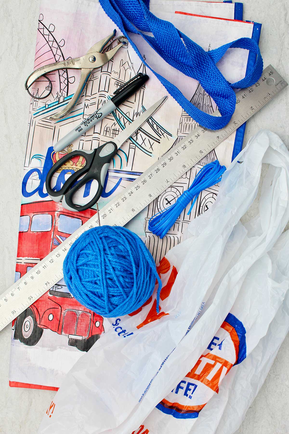 Supplies for a reusable bag sit upon cushion. Blue thread, reusable shopping bag, plastic shopping bags, ruler, sharpie, scissors and hole punch.