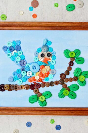 Completed bird button painting in wooden frame with scattered buttons around it.