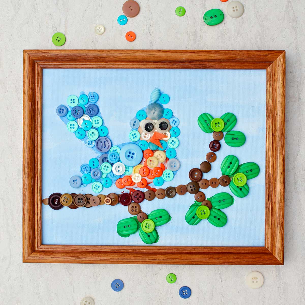 Completed bird button painting in wooden frame with scattered buttons around it.