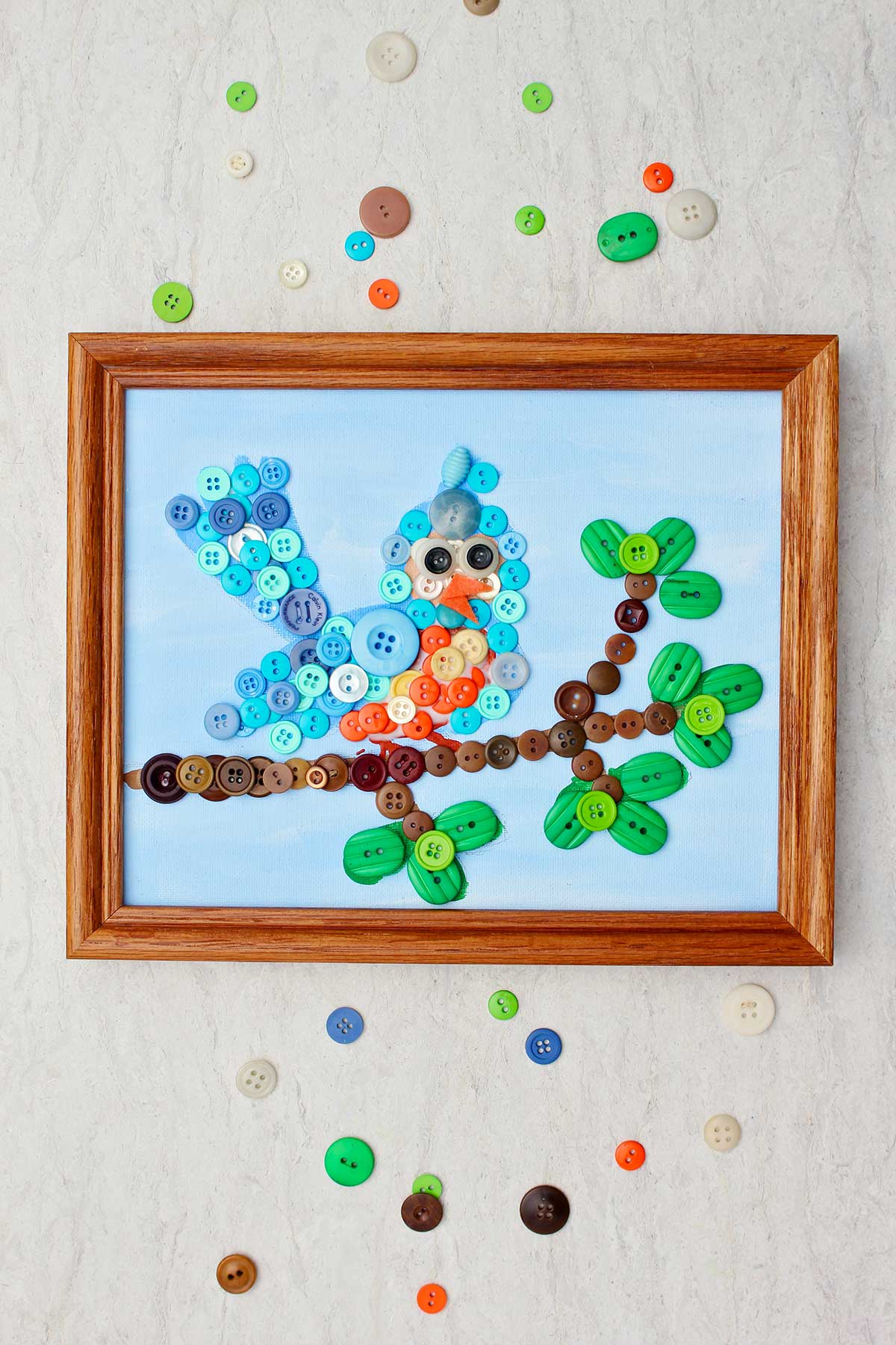 Completed bird button painting in wooden frame with scattered buttons around it.