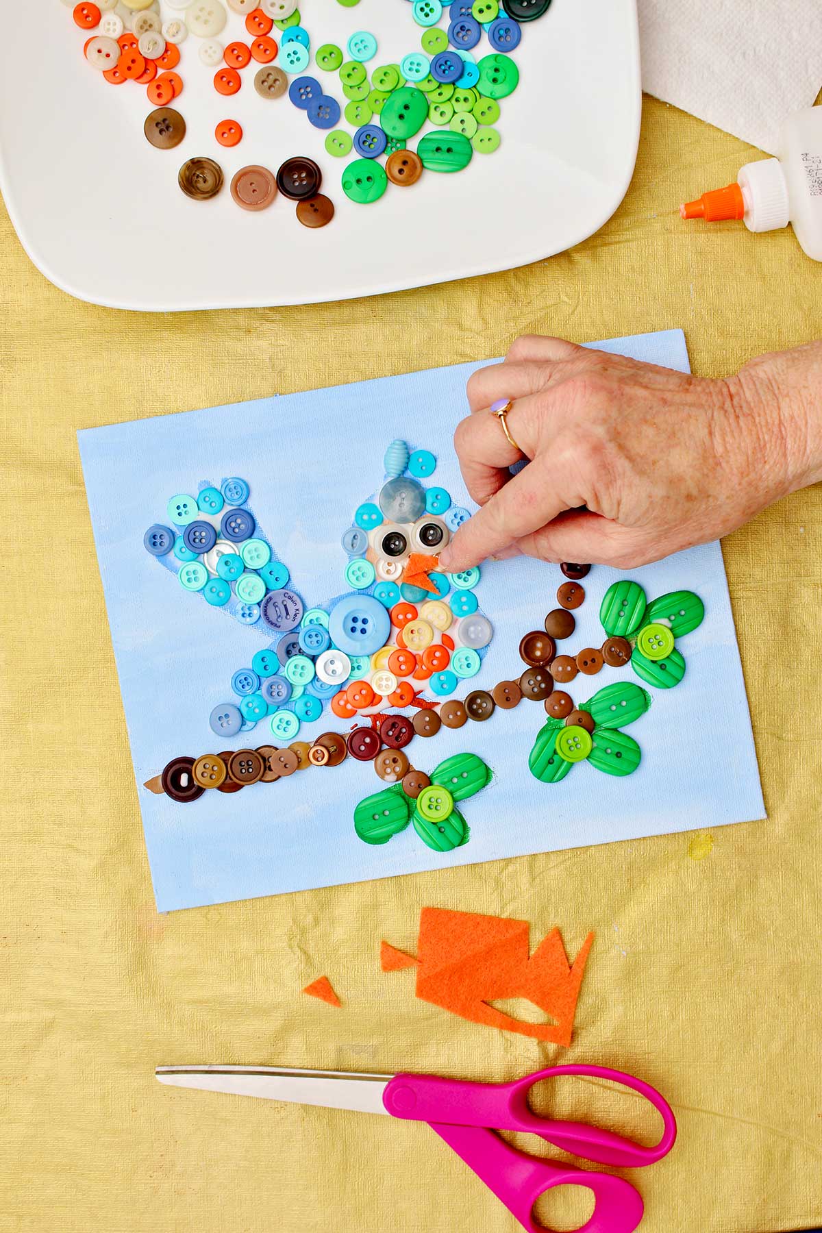 Person glueing down orange felt on bird for the beak in button craft project with other supplies near by.