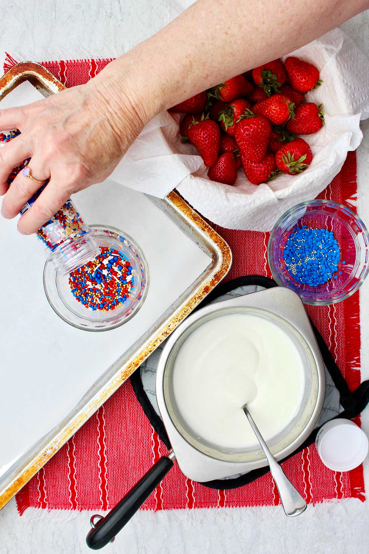 Person pouring sprinkles into glass bowl sitting on parchment covered cookie sheet.