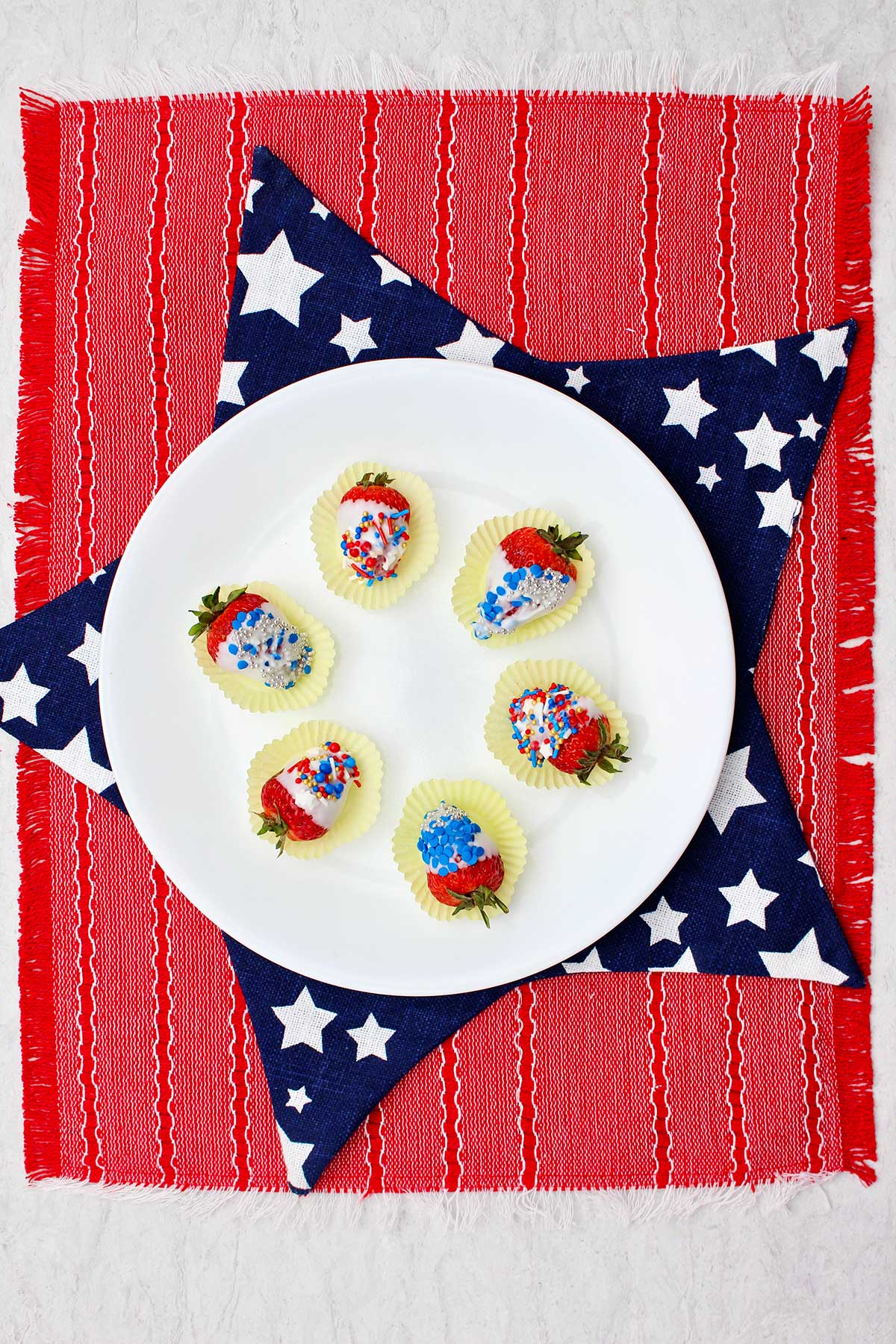 White plate of completed white chocolate dipped strawberries sitting on a blue and white star mat and red placemat.