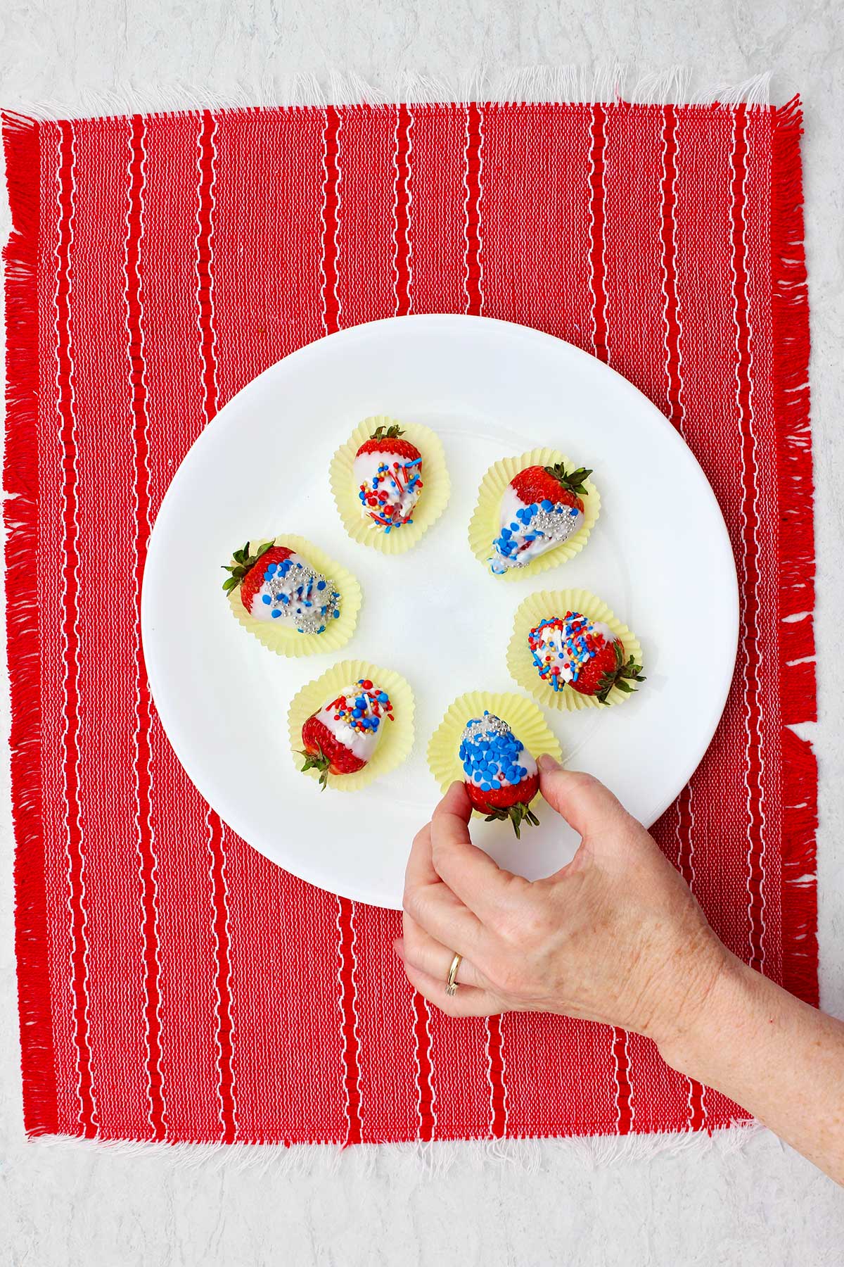 Person picking up a white chocolate dipped strawberry from a plate sitting on a red placemat.