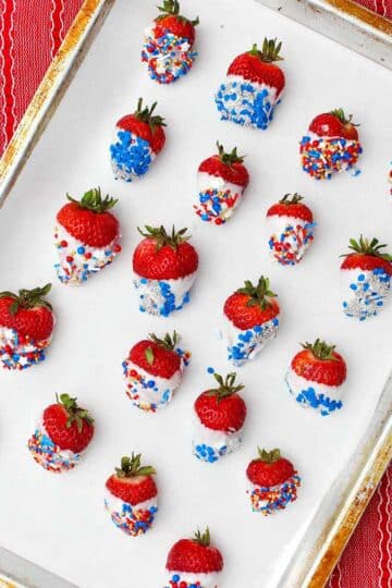 Completed white chocolate dipped strawberries with blue, red and silver sprinkles sitting on a cookie sheet with parchment paper.