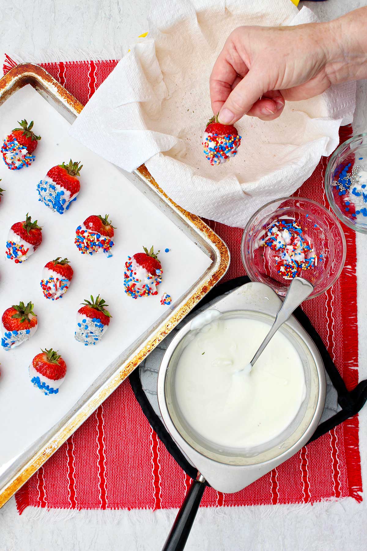 Hand holding completed white chocolate dipped strawberry near warming almond bark and cookie sheet with completed chocolate dipped strawberries.