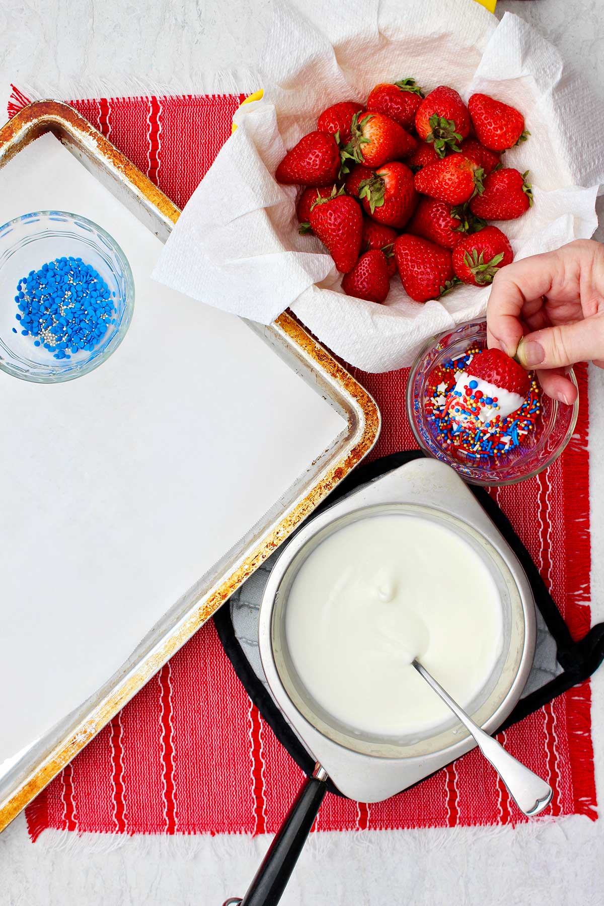 Hand holding white chocolate dipped strawberry over a bowl of sprinkles near warming almond bark and cookie sheet with parchment paper.