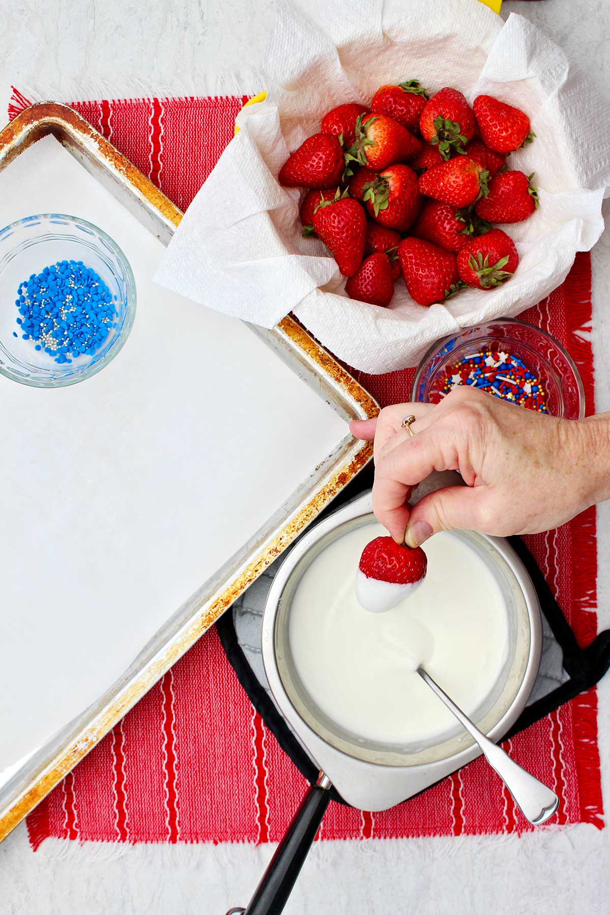 Person dipping strawberry in warming almond bark near bowl of strawberries, bowl of sprinkles and cookie sheet with parchment paper.