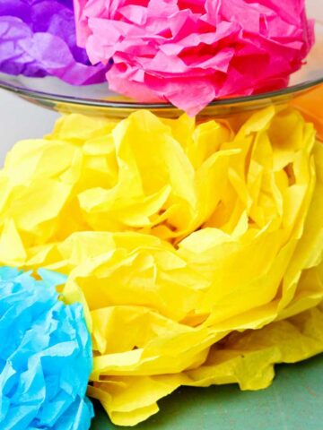 Completed colorful tissue paper flowers, a purple and pink one resting on a glass cake stand with others underneath.