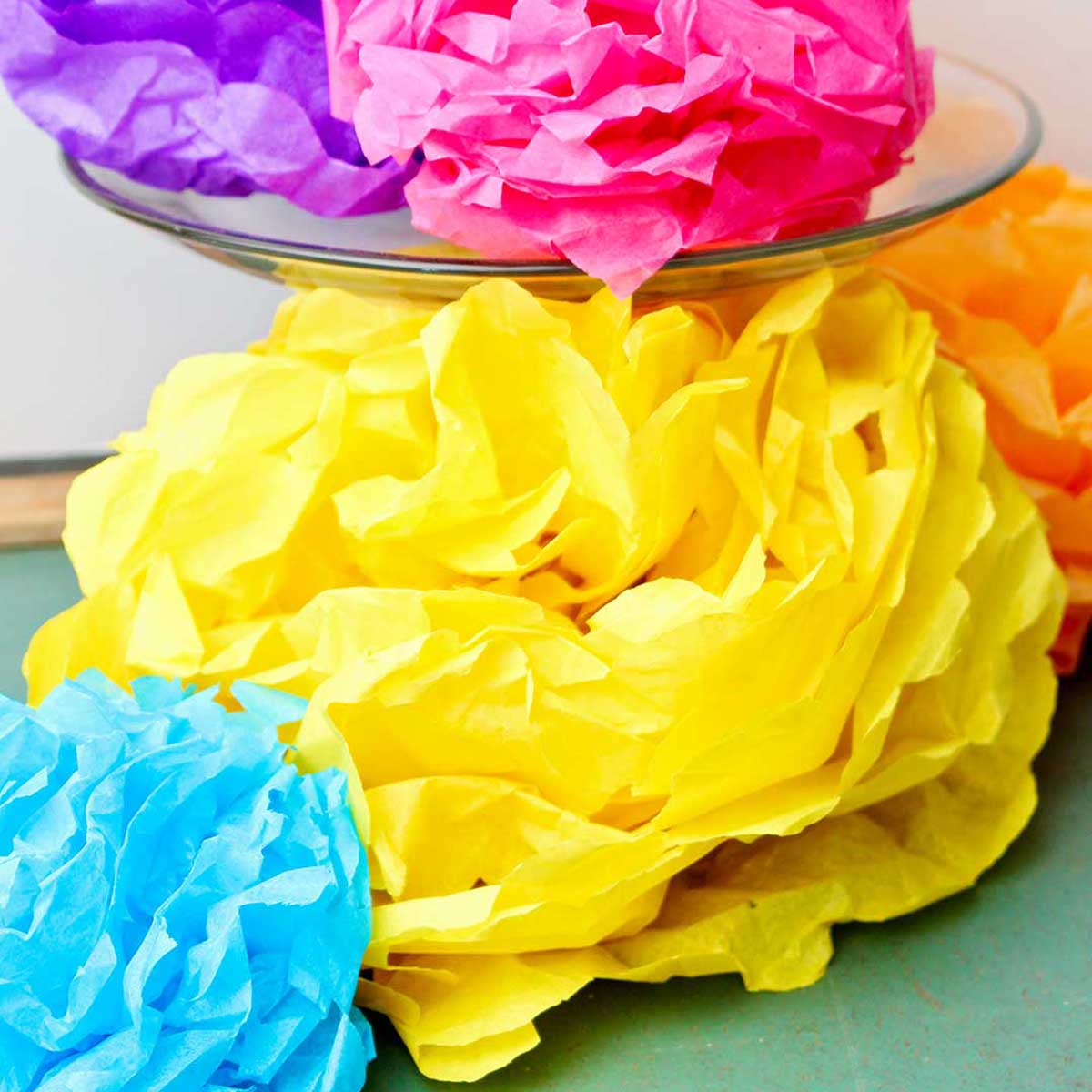 Completed colorful tissue paper flowers, a purple and pink one resting on a glass cake stand with others underneath.