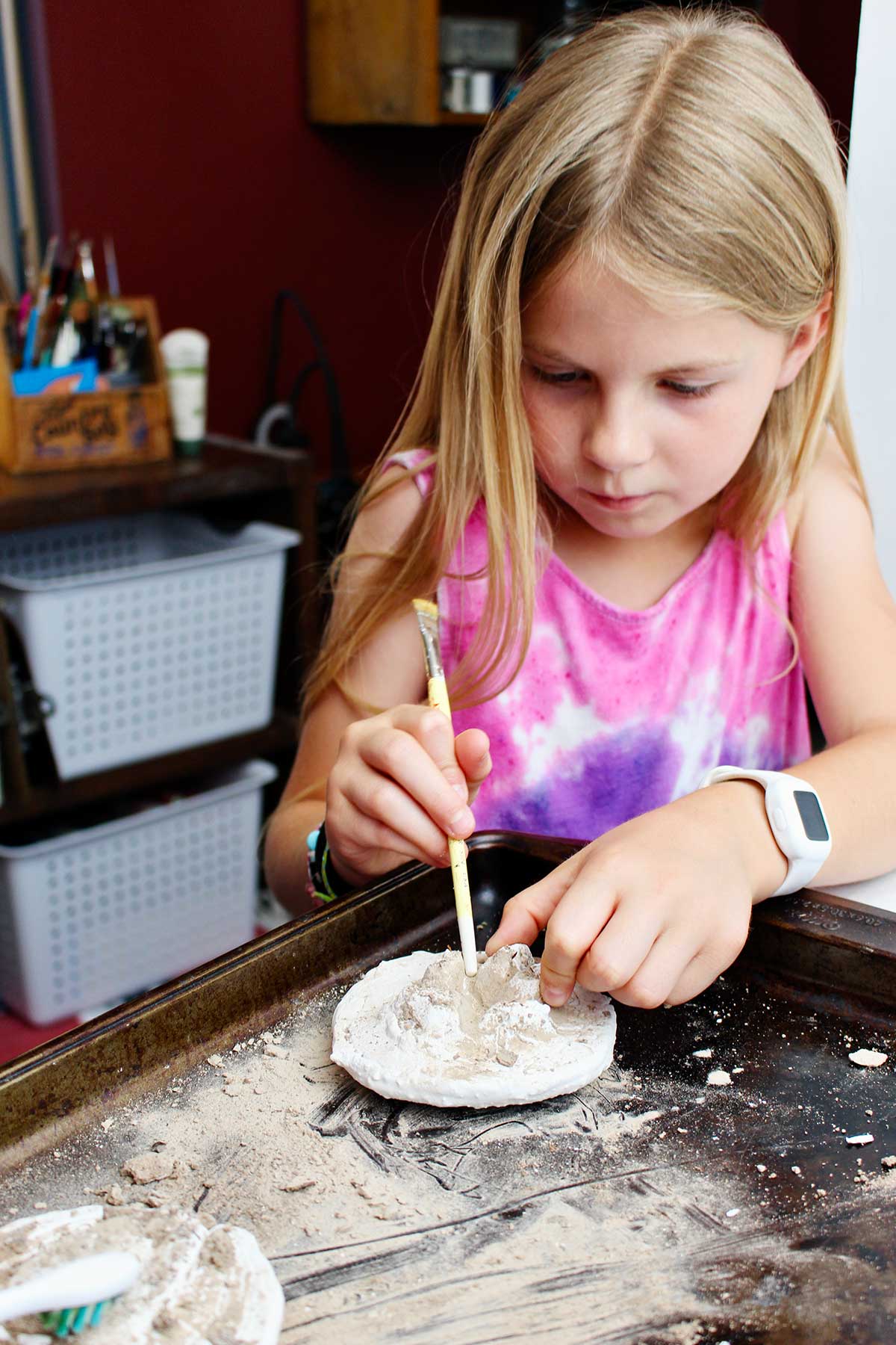 Young girl with blonde hair using end of paintbrush to get dirt out of her plaster cast of an animal track.