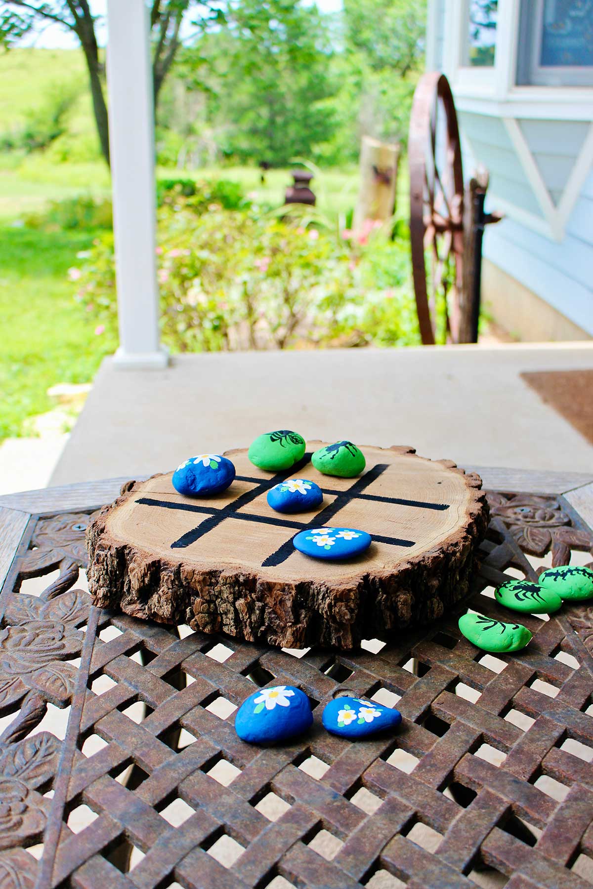 Wooden tic tac toe board with painted rocks on an iron table outside.