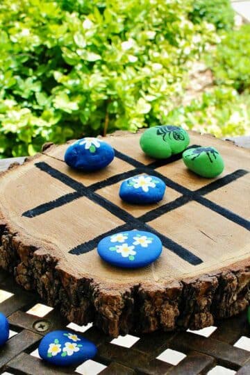 Wooden tic tac toe board with painted rocks on an iron table outside.