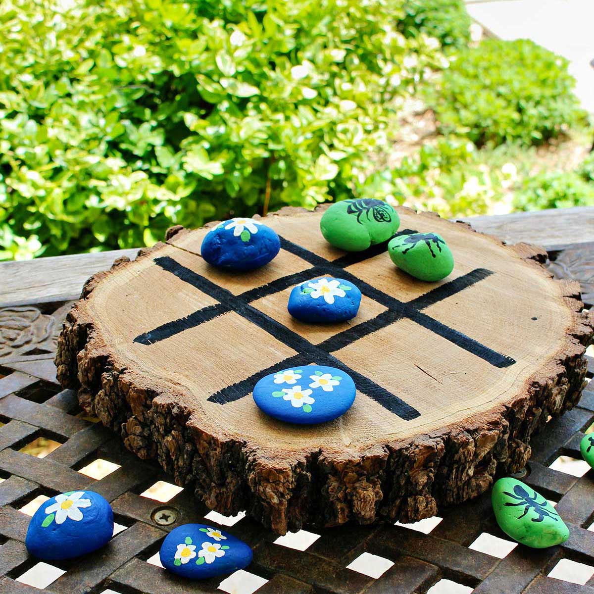 Wooden tic tac toe board with painted rocks on an iron table outside.