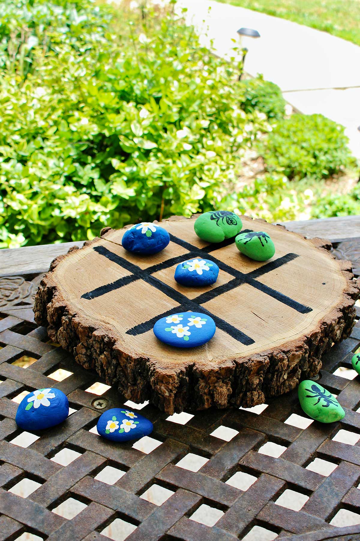 Wooden tic tac toe board with painted rocks on an iron table outside.