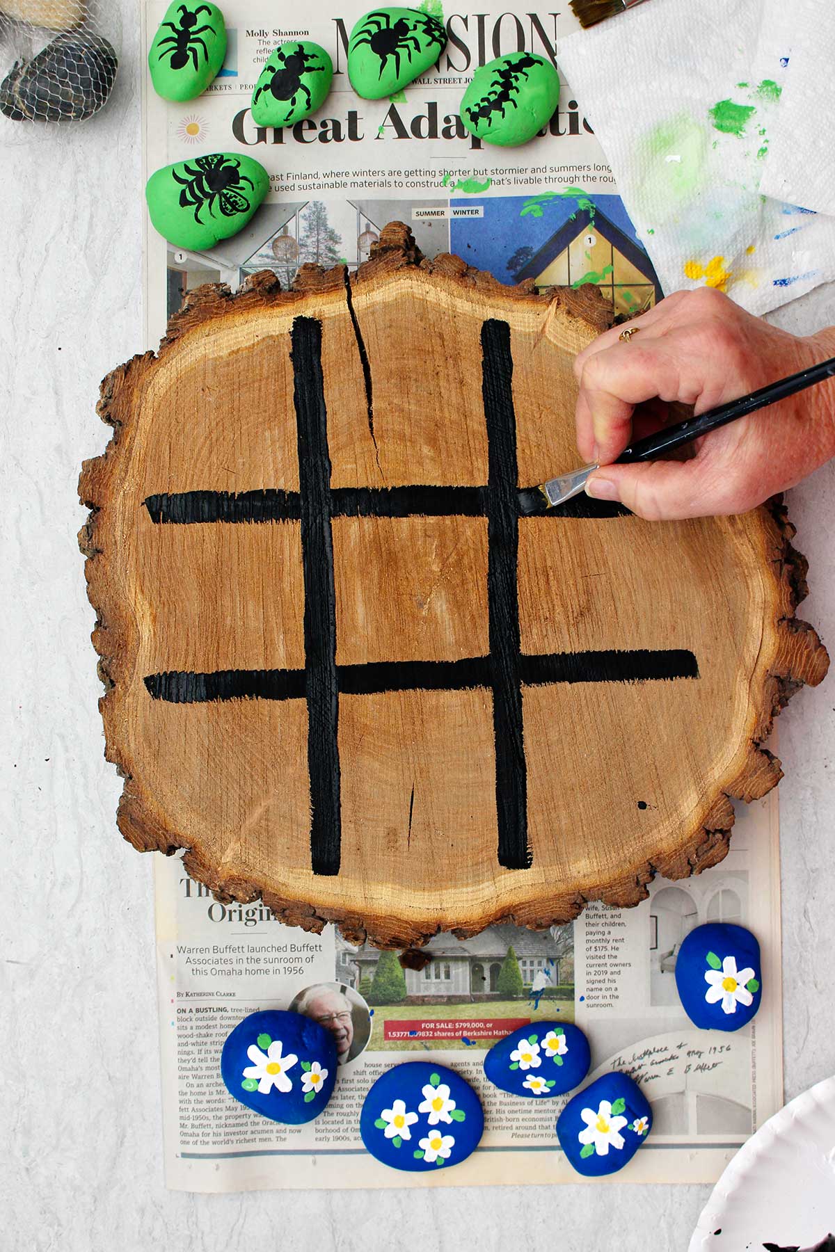 Person painting black lines on a circular cut of wood to make tic tac toe board.