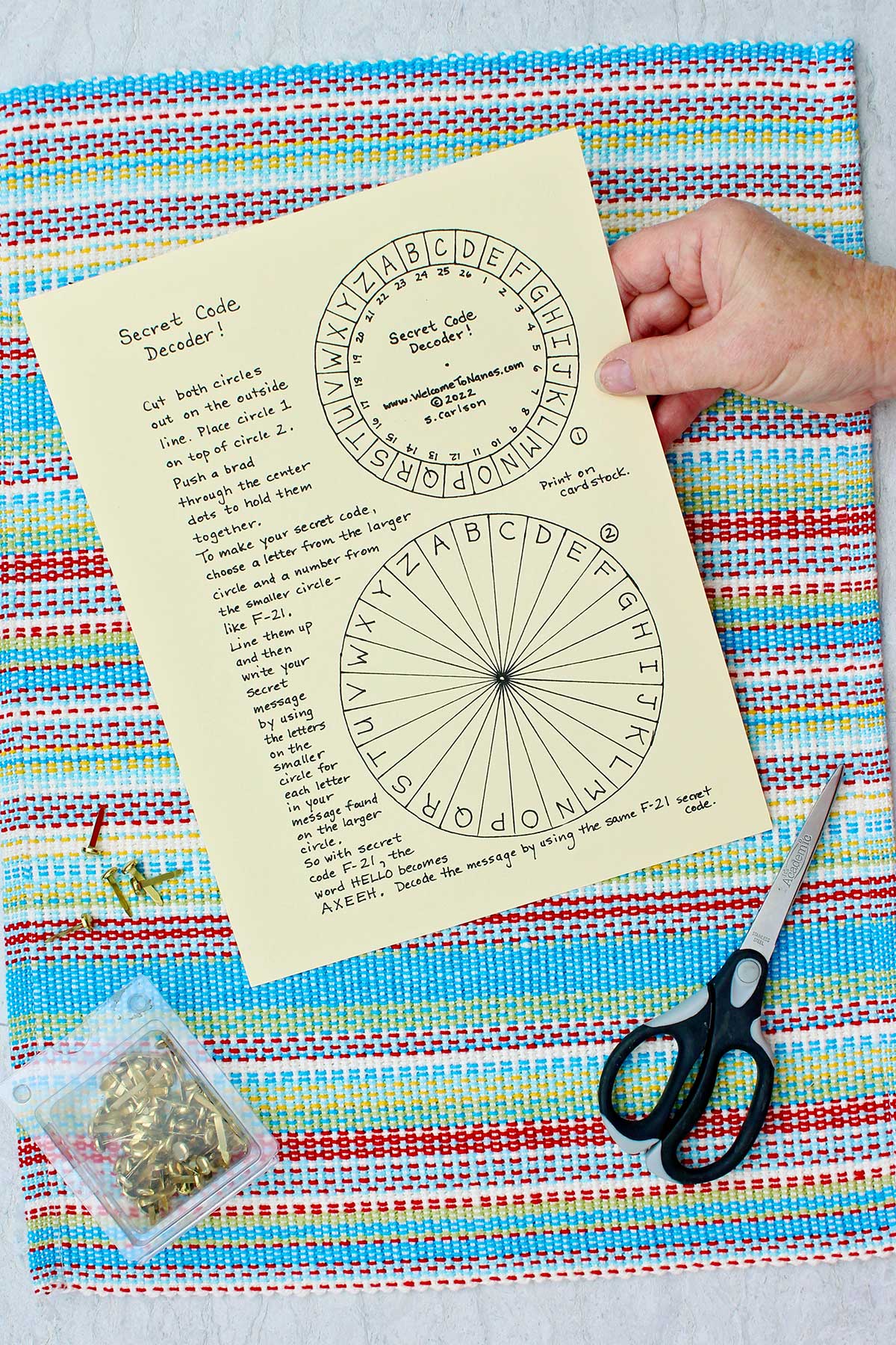 Person holding secret message decoder printout with brass brads and scissors resting on a colorful place mat.