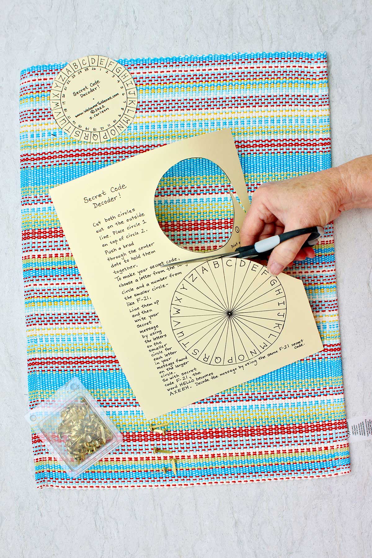 Person cutting out Secret Message Decoder Wheel from printout with brass brads near by against a multicolored placemat.