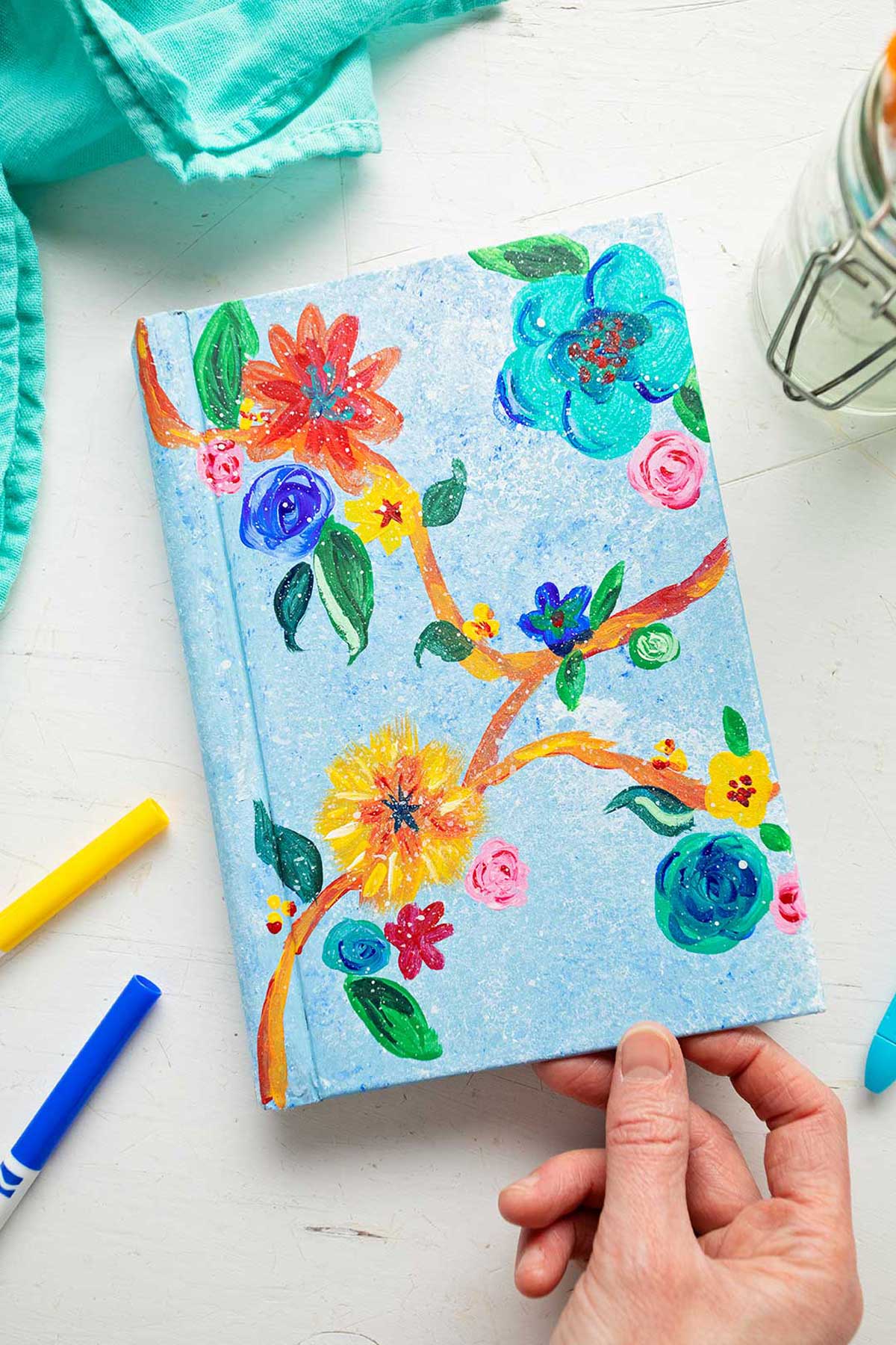 Hand holding bottom of painted book cover with brightly colored flowers and leaves.