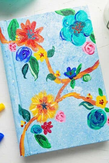 Painted book cover with brightly colored flowers and leaves near markers and a teal linen.