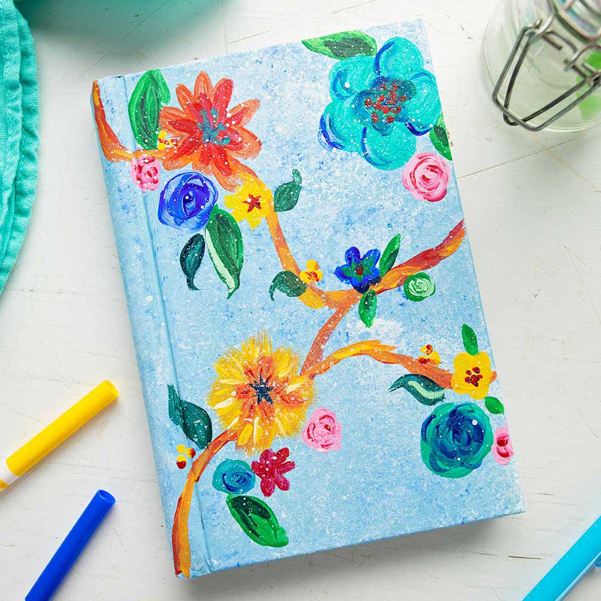 Painted book cover with brightly colored flowers and leaves near markers and a teal linen.