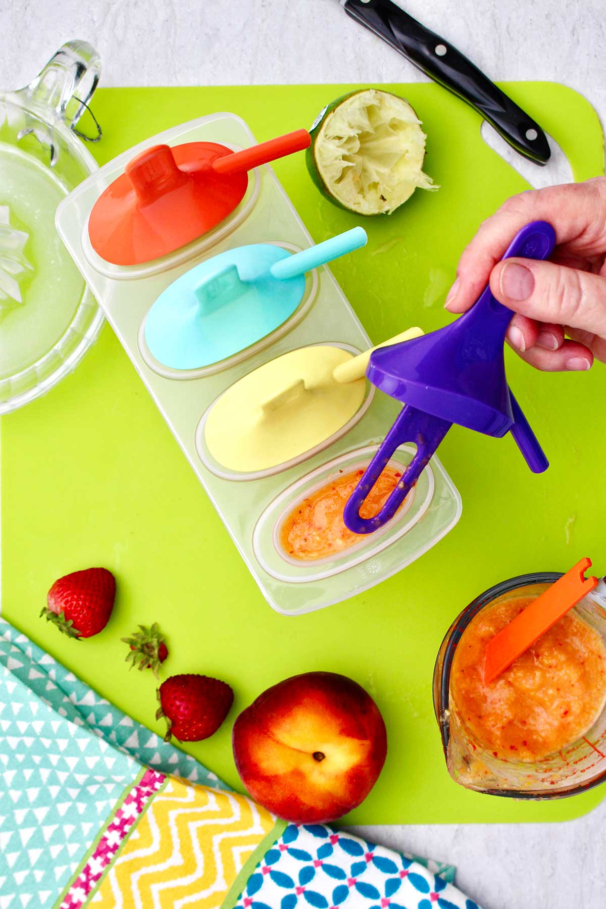 Person placing a purple popsicle holder inside popsicle mold with other fruits and supplies near by.