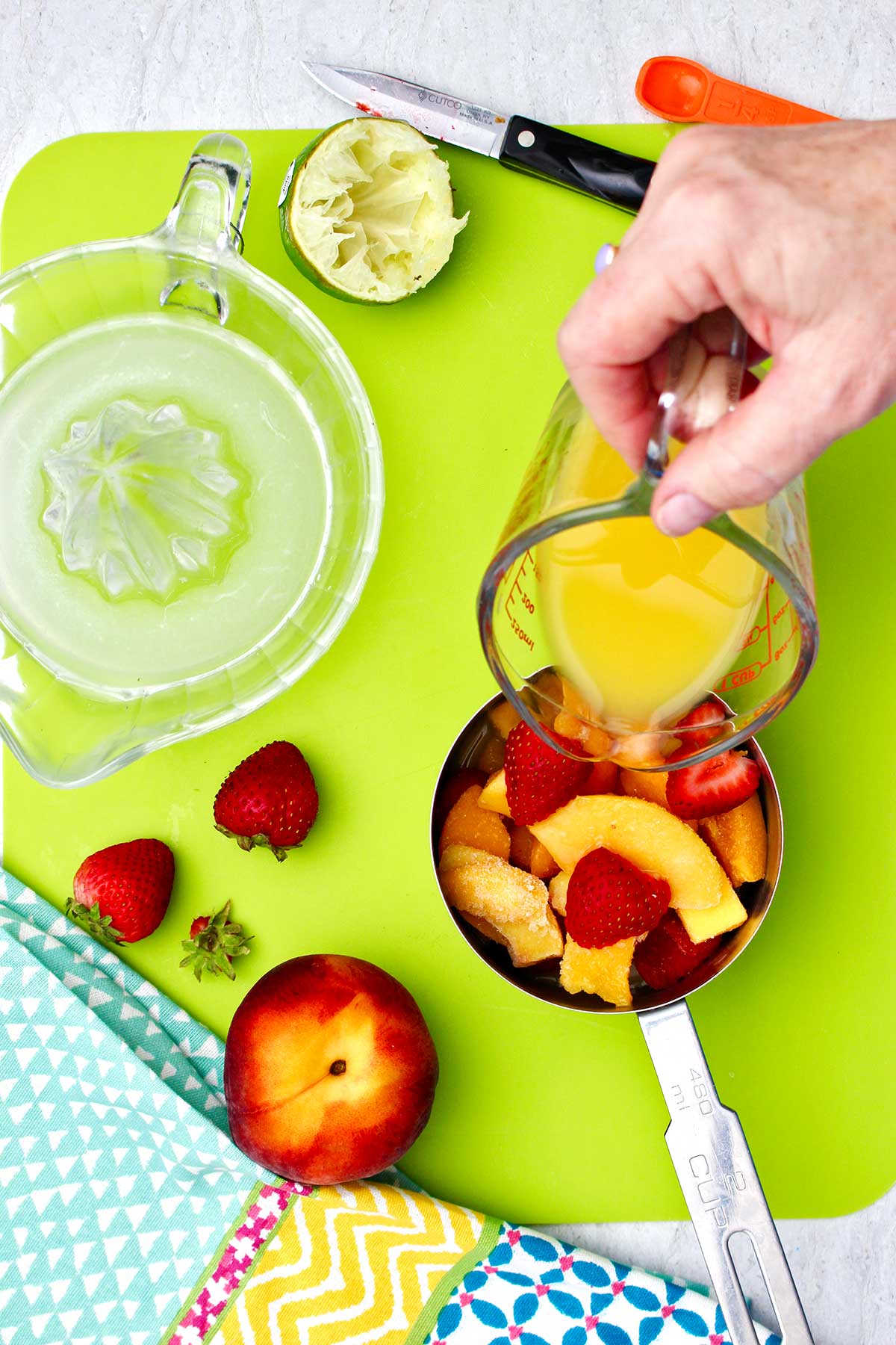 Person pouring orange juice into a measuring cup of assorted frozen fruit on a bright green cutting board.